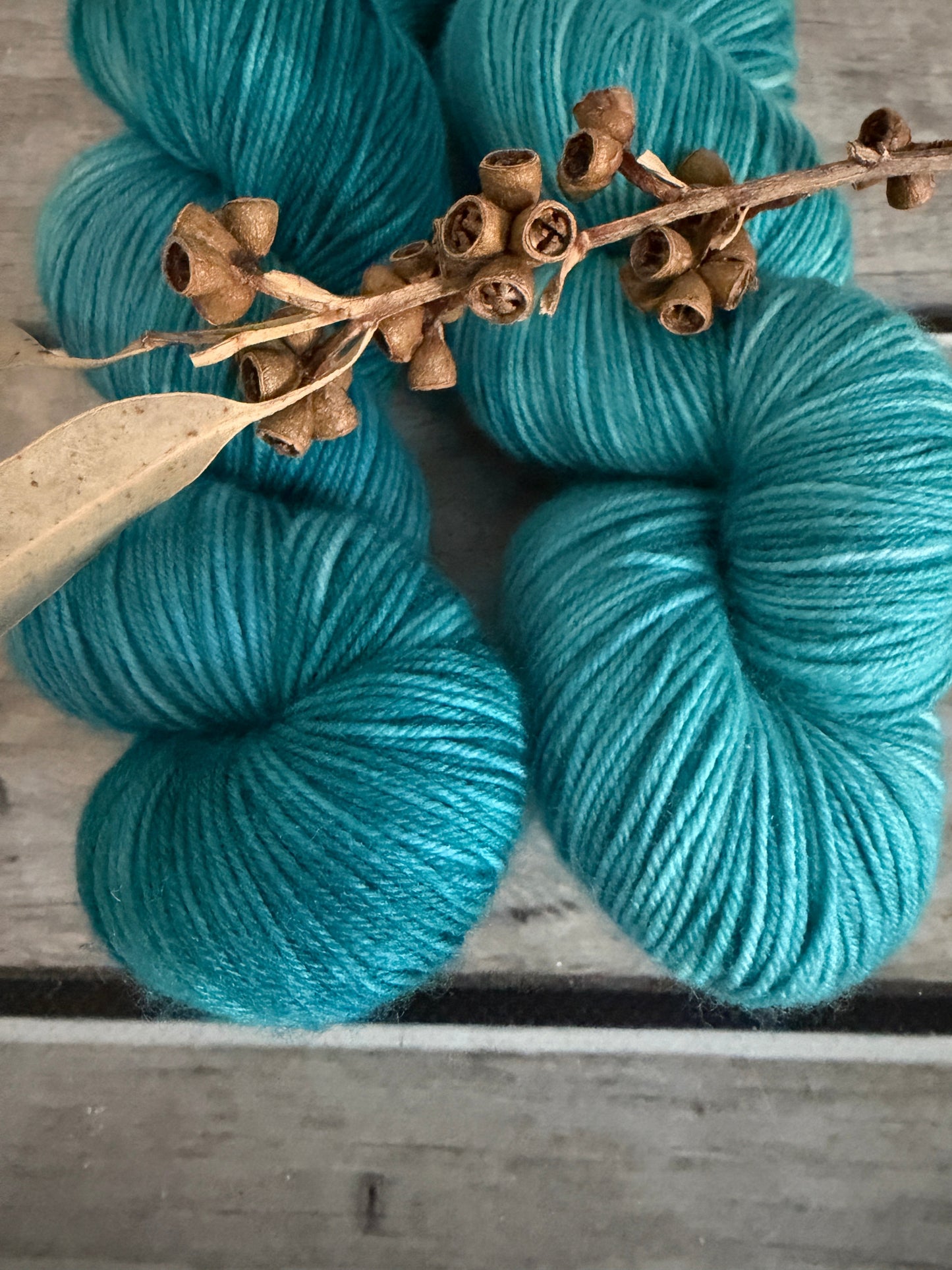 Two skeins of turquoise blue yarn arranged on a weathered gray wooden surface, with a dried eucalyptus branch placed diagonally across the foreground.