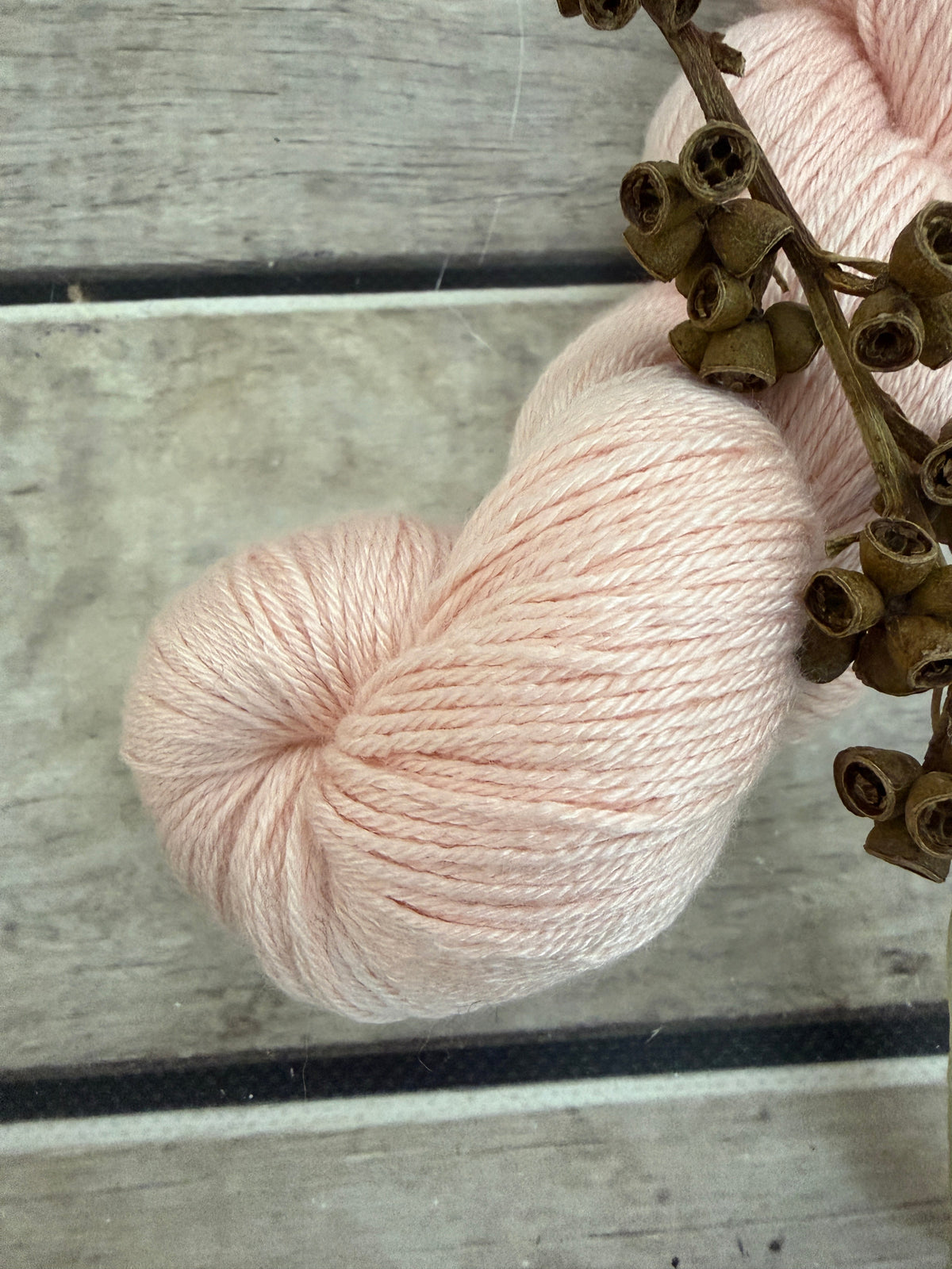 A close-up of a pale blush pink skein of yarn resting on a rustic gray wooden surface, with dried brown buds in the foreground.