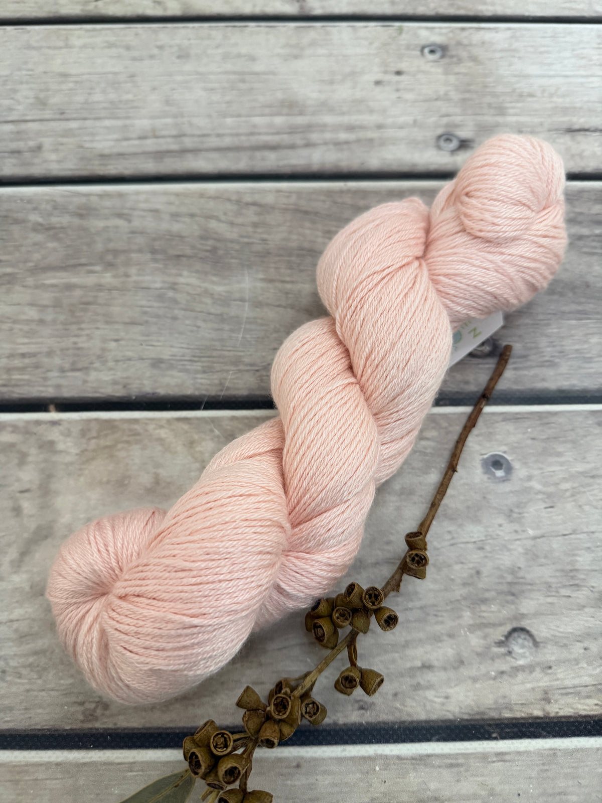 A single skein of soft blush pink yarn arranged diagonally on a weathered gray wooden surface, with a dried brown floral stem placed beside it.