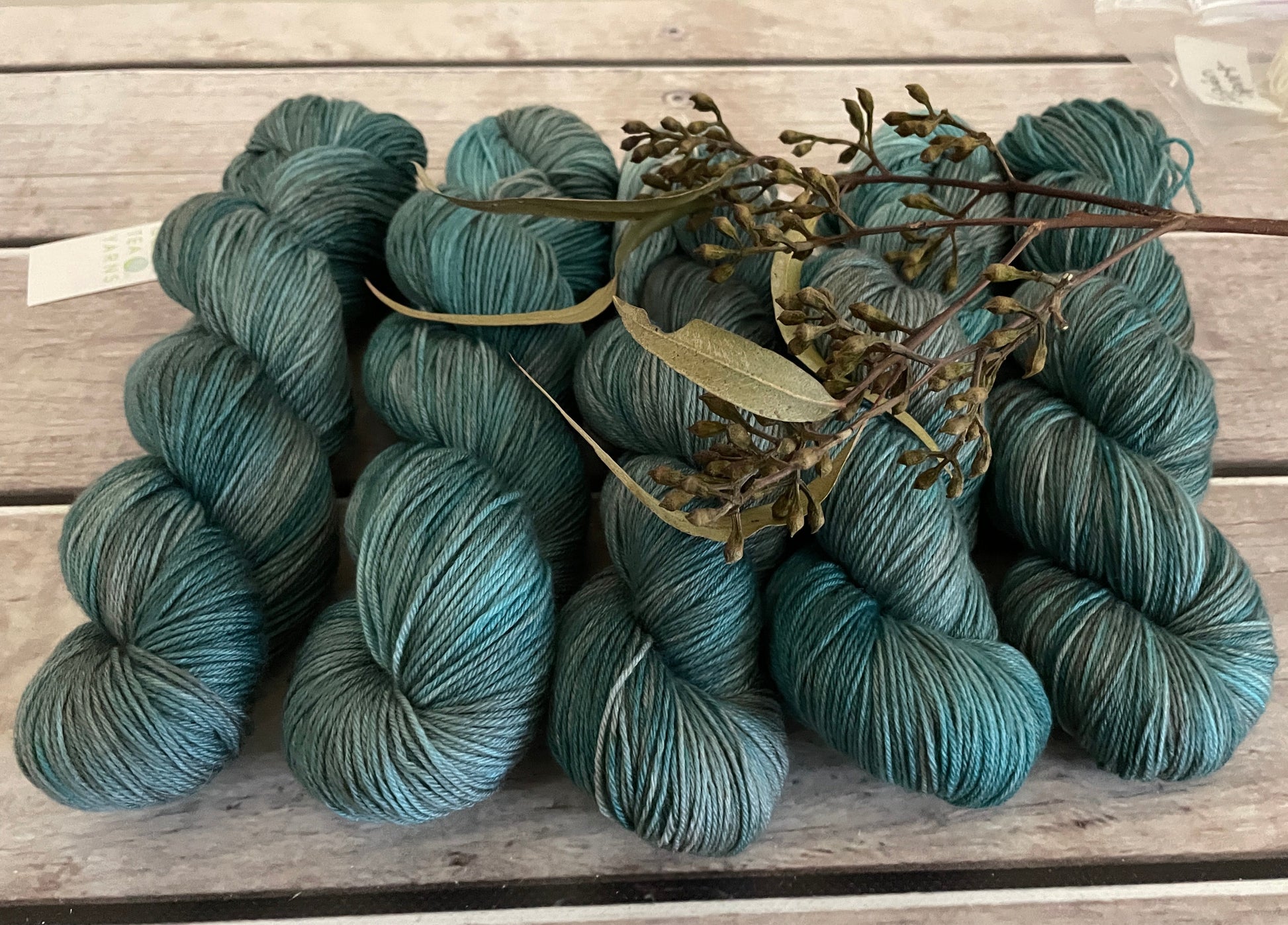Five skeins of blue green yarn sit on top of a wooden boards. They have a sprig of ecalyptus sitting on them