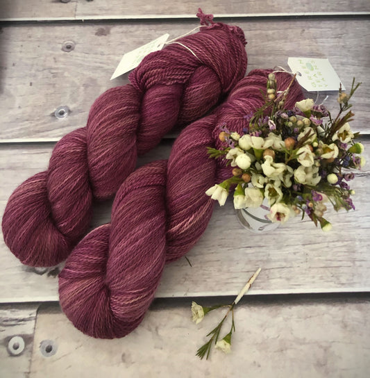 A skein of purple yarn in front of a floral arrangement, labeled 'White Cloud' and indicating it is a 3 ply silk and merino blend.